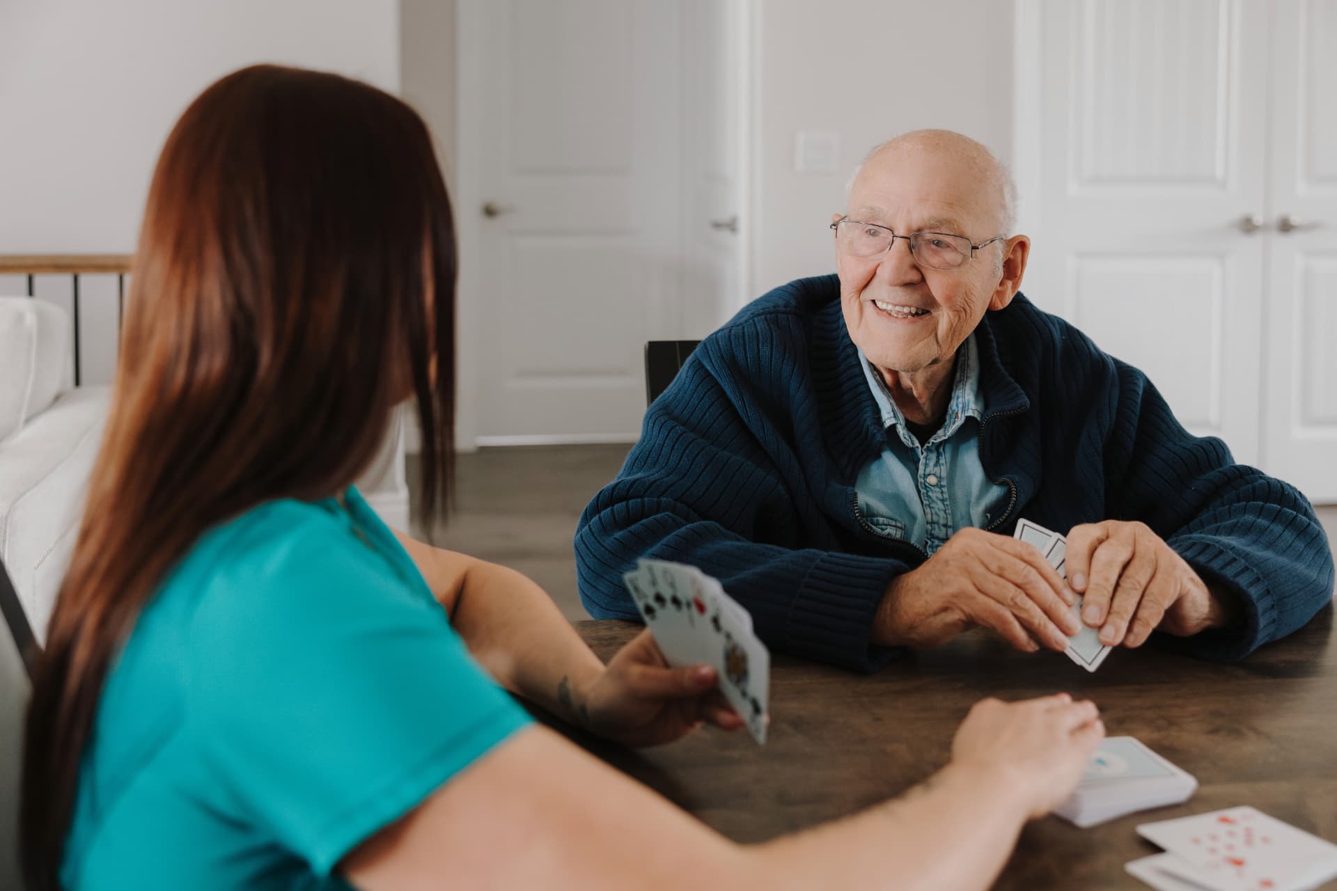 Caregiver in teal scrubs playing cards with a smiling senior man at a home table